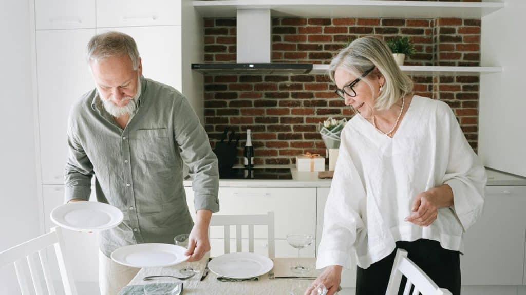 A senior couple setting the table together in a kitchen.
