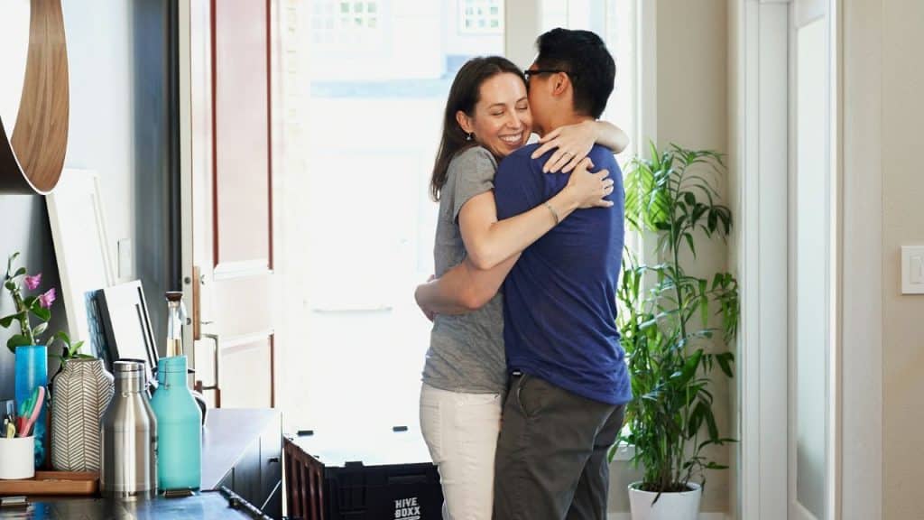 A woman smiles and hugs a man in a blue shirt in an entryway.