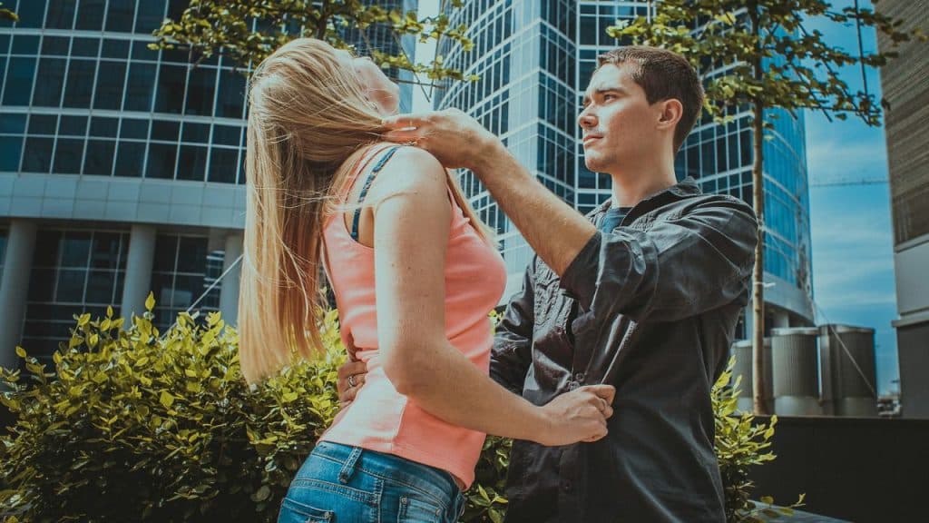 A man in a dark shirt reaches toward a woman’s neck while standing outdoors near a building.