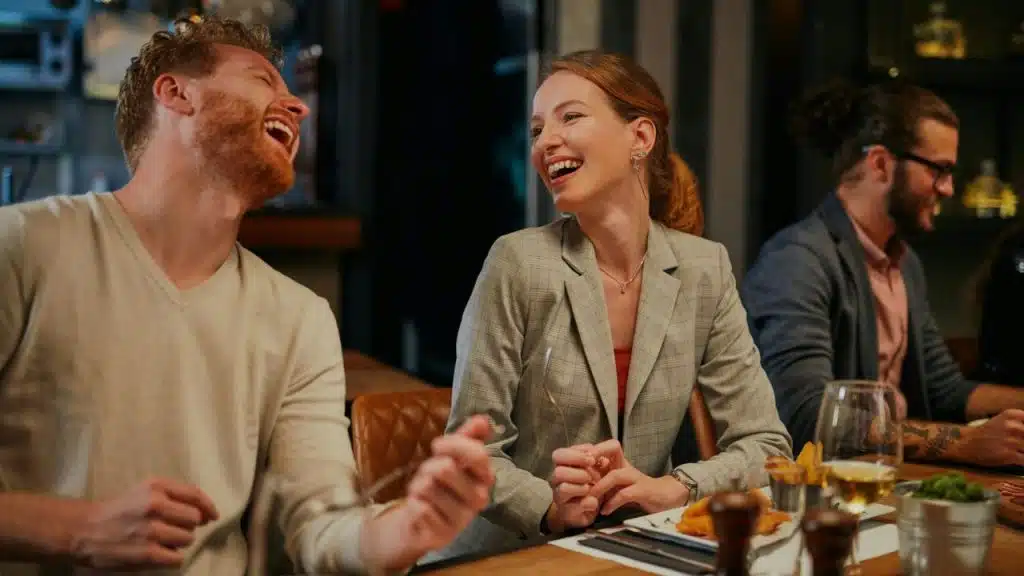 A man and woman in business casual attire laugh together while seated at a dinner table.