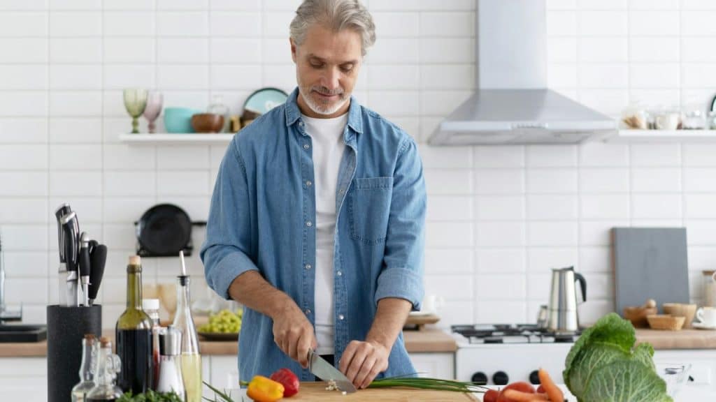 A man with grey hair chops green onions on a wooden board in a white kitchen.