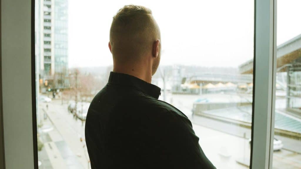 A man in a black shirt is seen from behind, looking out a large window.