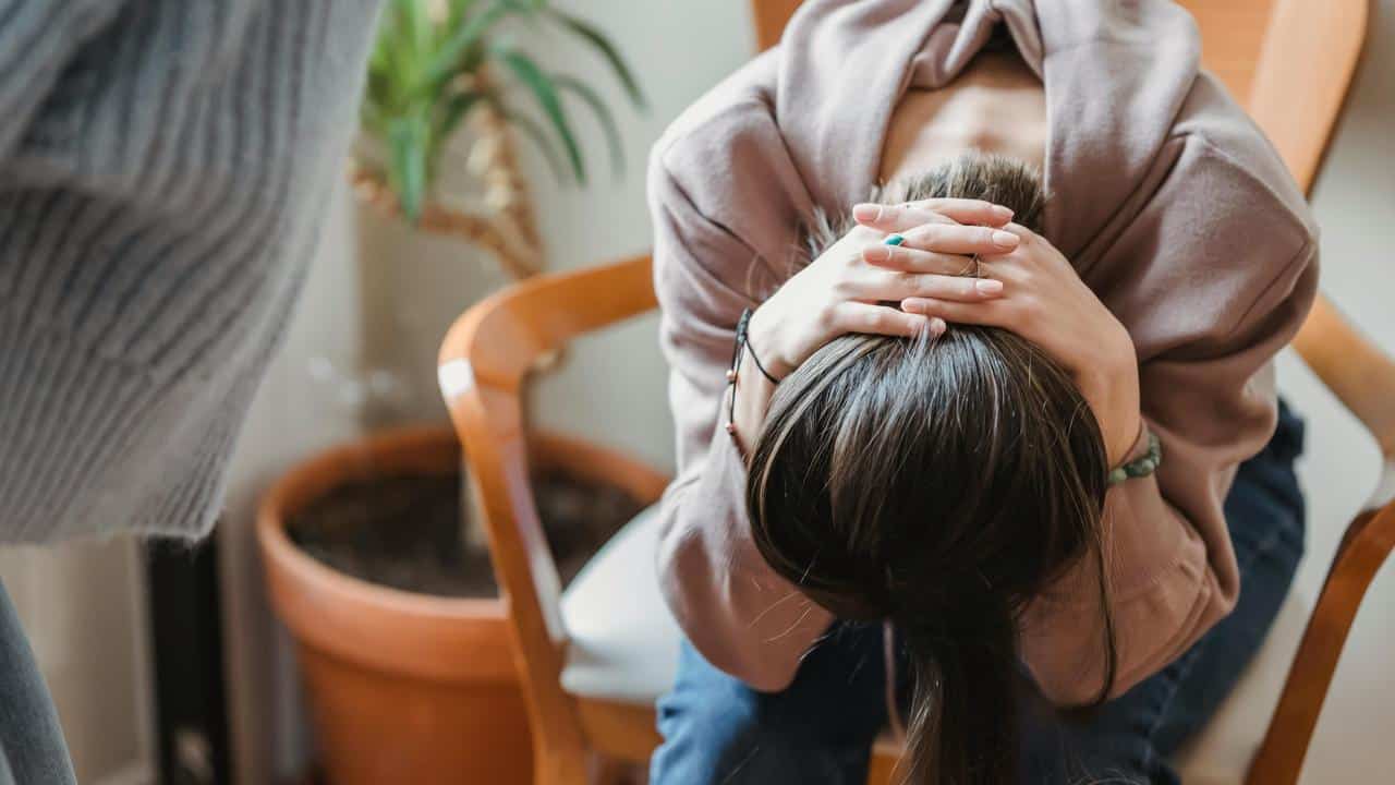 A person sitting on a chair with their head bent forward and hands held over their hair.