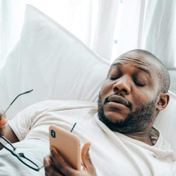 A man reclining in bed while holding a phone and glasses.