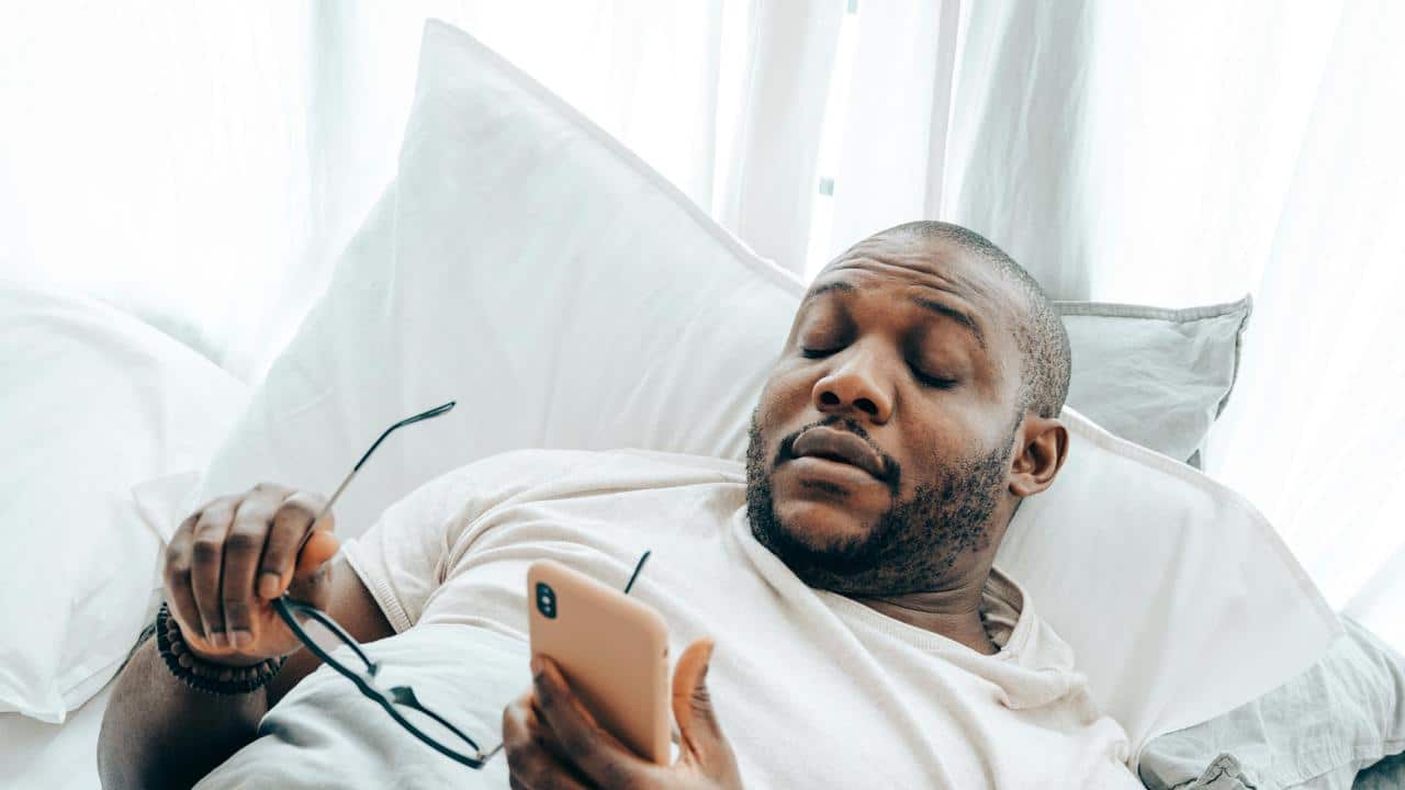 A man reclining in bed while holding a phone and glasses.