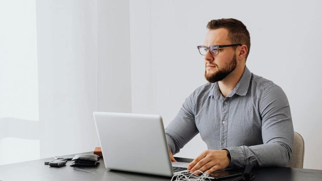 A man wearing glasses working on a laptop at a desk in a bright room.