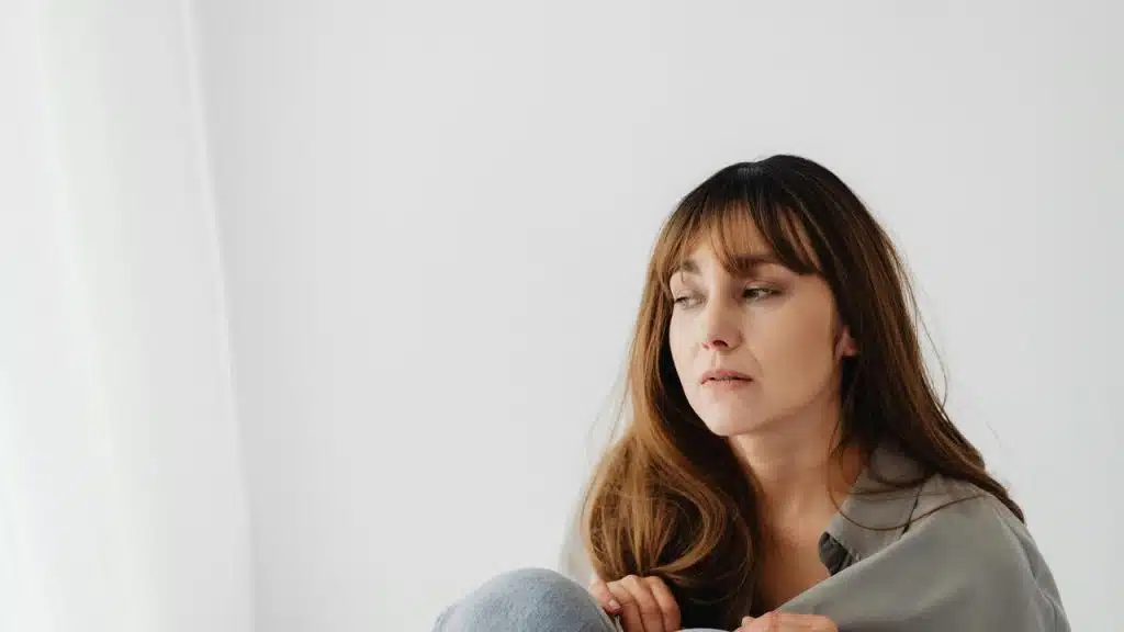 A woman sitting indoors and gazing thoughtfully toward a bright window.