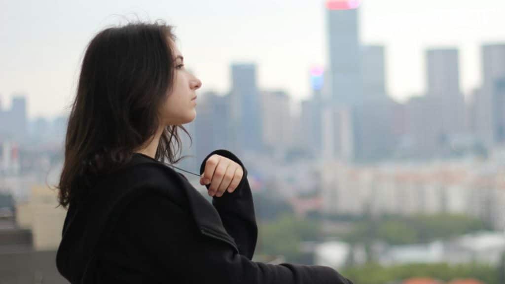 A woman in a black jacket looks out toward a blurred cityscape in the distance.