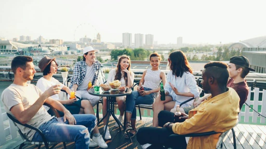 A diverse group of people sits around a table on a rooftop, talking and holding drinks.