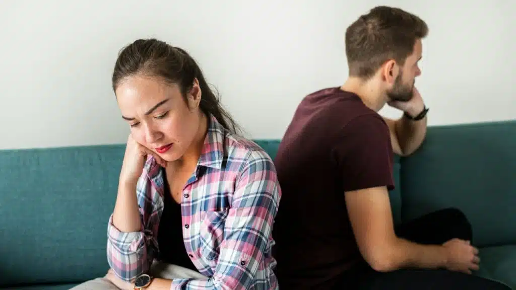 A man and woman sit back-to-back on a teal sofa looking away from each other.