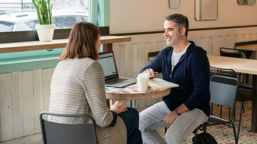A man and woman talk at a small cafe table with a laptop and coffee.