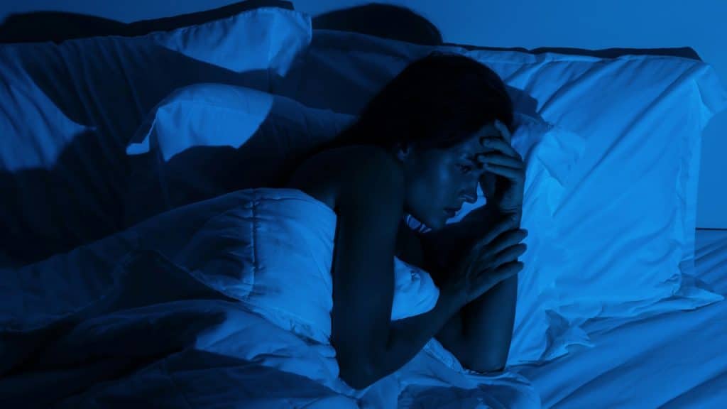 A woman lying awake in bed in the dark, clutching her head.