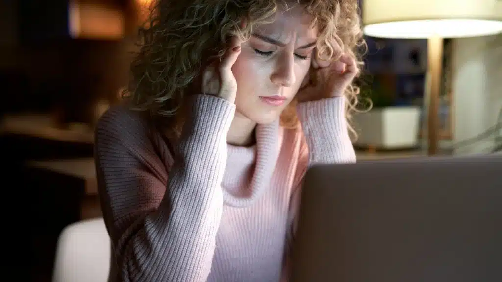 A woman sitting in front of a laptop, holding her temples with her eyes closed.