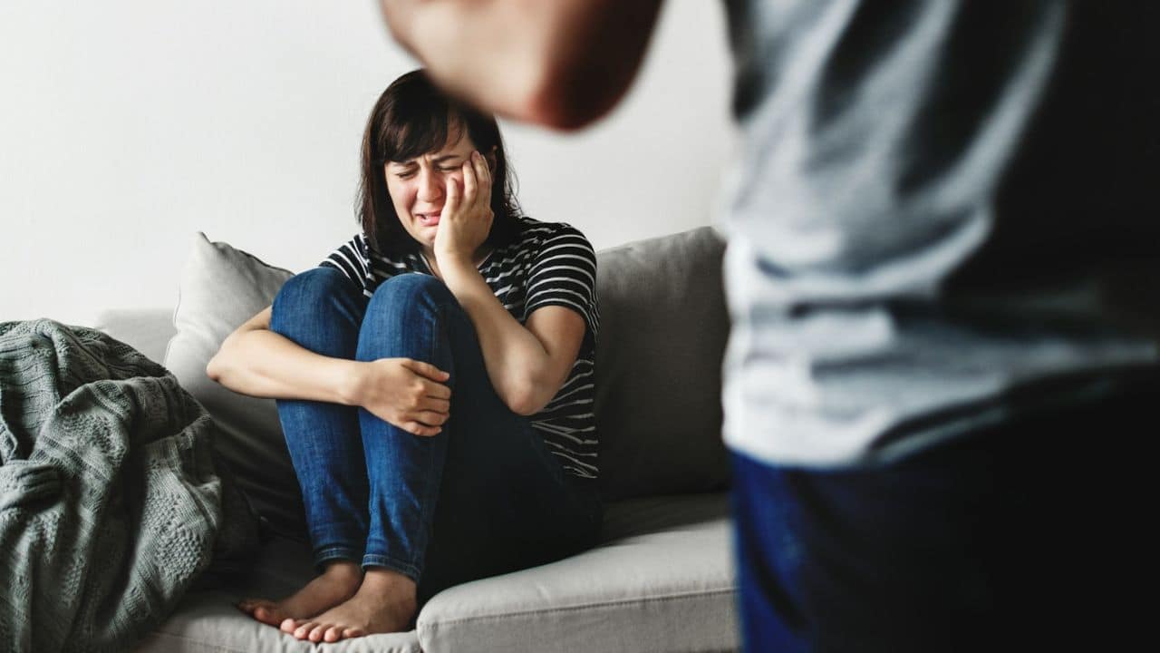 Distressed woman crying on a couch, covering her face, with a man standing nearby.