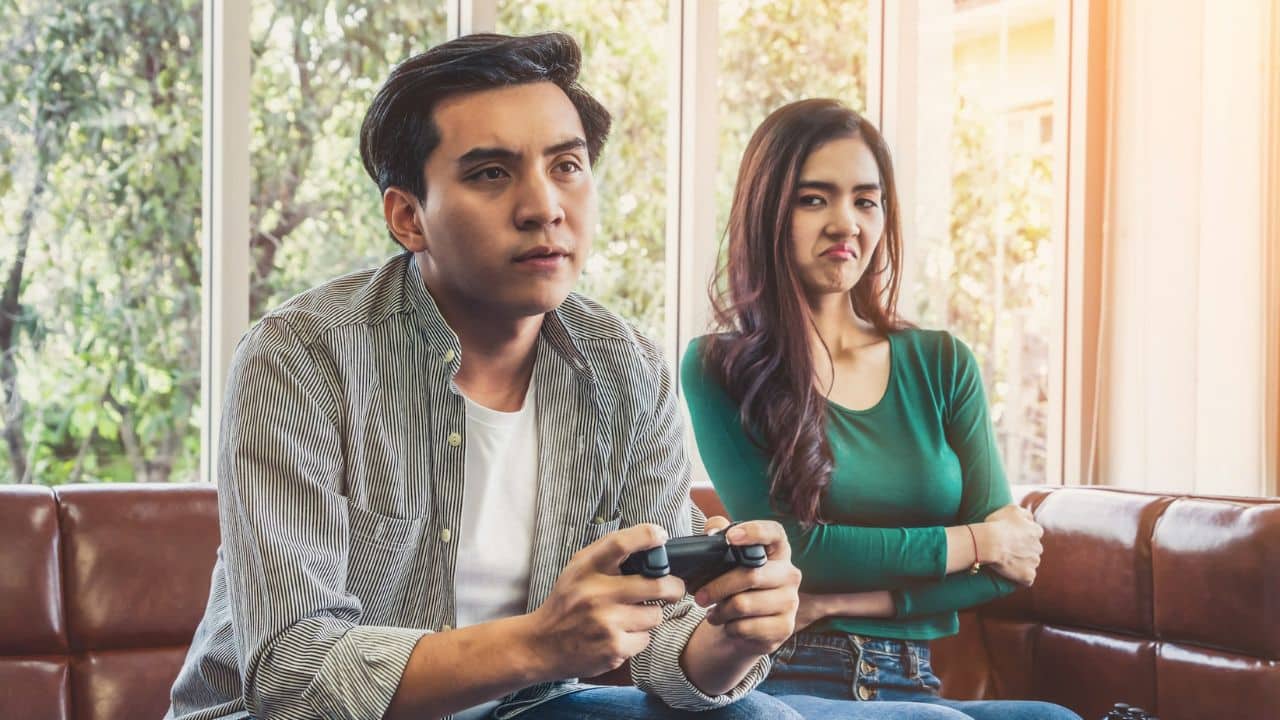 A man plays a video game on a couch while a woman next to him gives a disapproving look.
