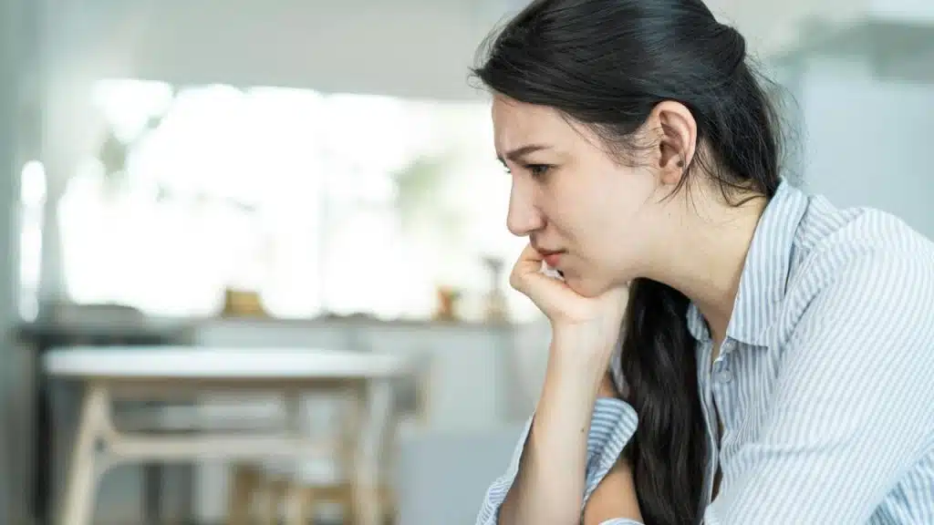 A woman with dark hair sits indoors, looking down with a pained expression and her chin resting on her hand.