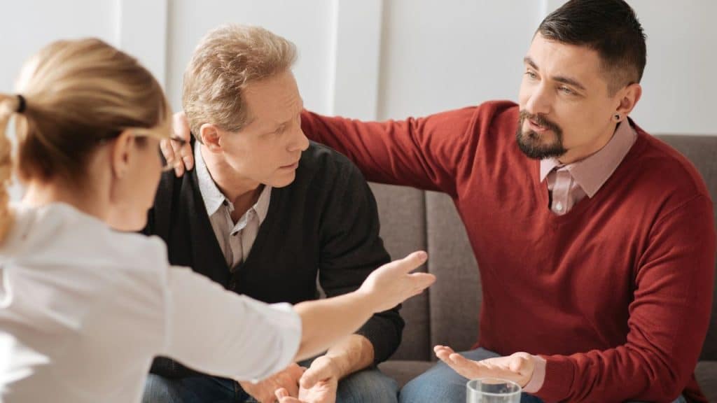 Three adults engage in a serious conversation; a man comforts another while a third gestures.