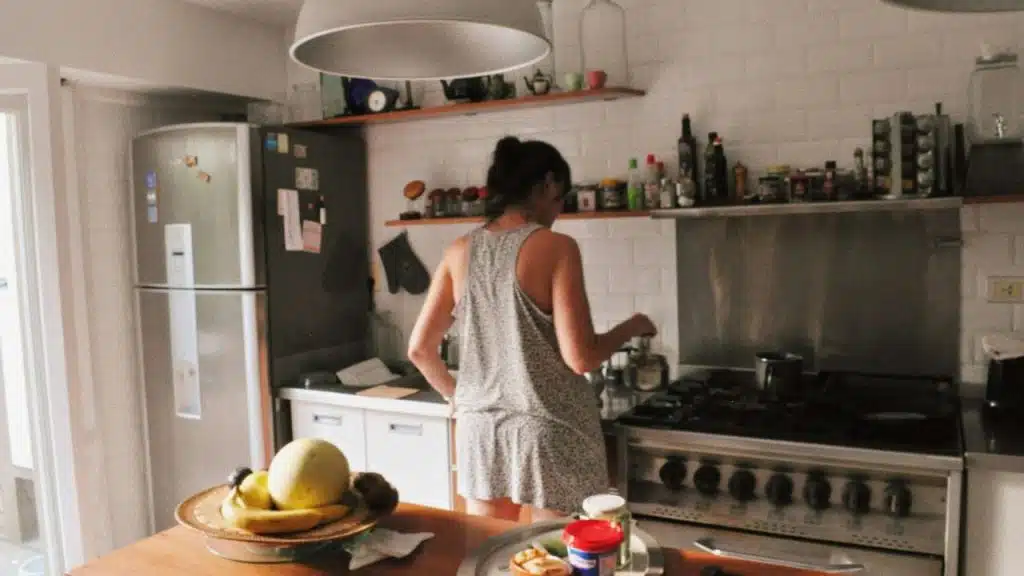 Woman in a sleeveless top cooking on a large stove in a kitchen with white tile walls.
