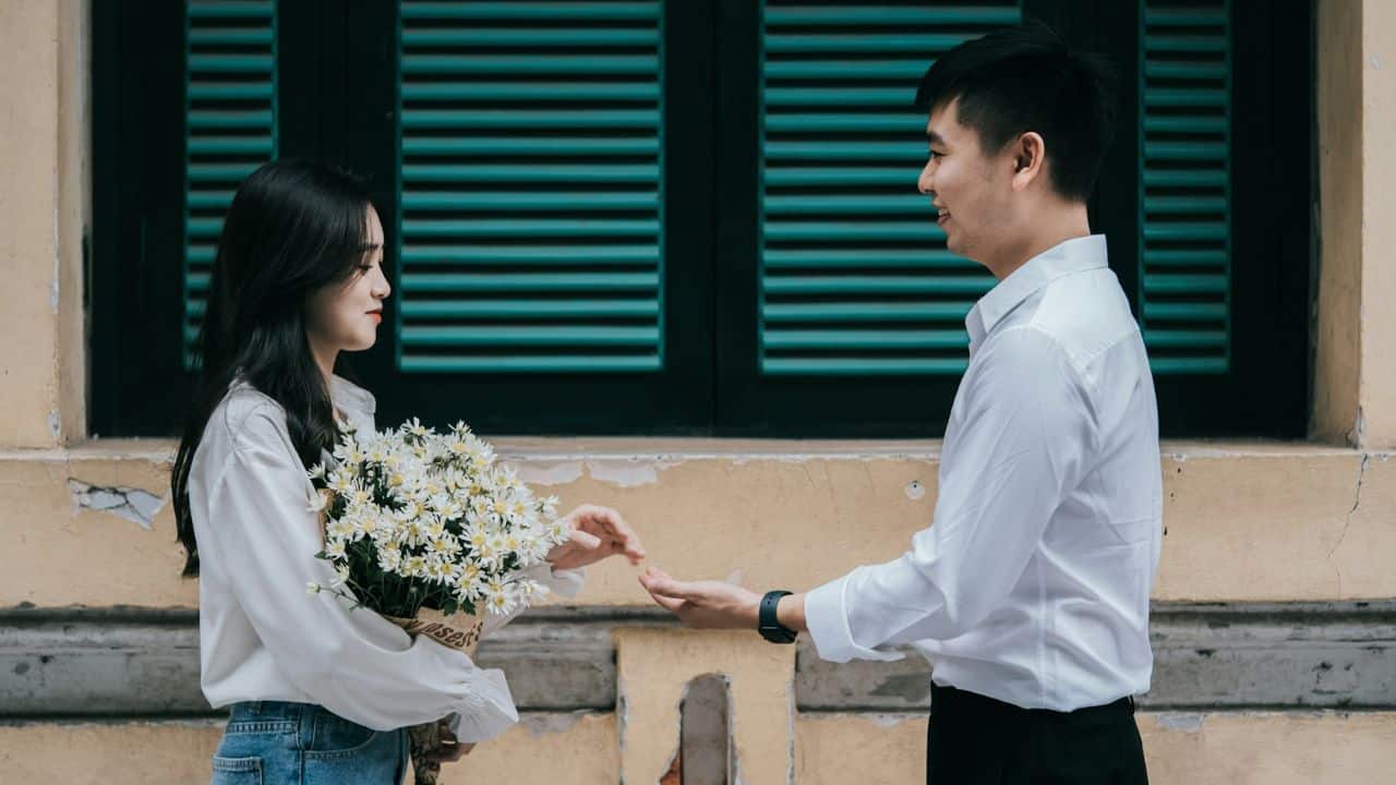 A man and woman in white shirts stand facing each other; the woman holds white flowers.