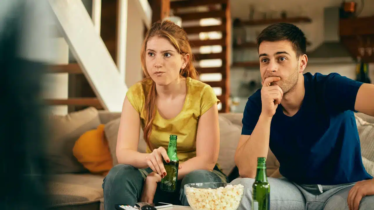 A couple intently watching TV on a couch, holding beers with popcorn on the coffee table.