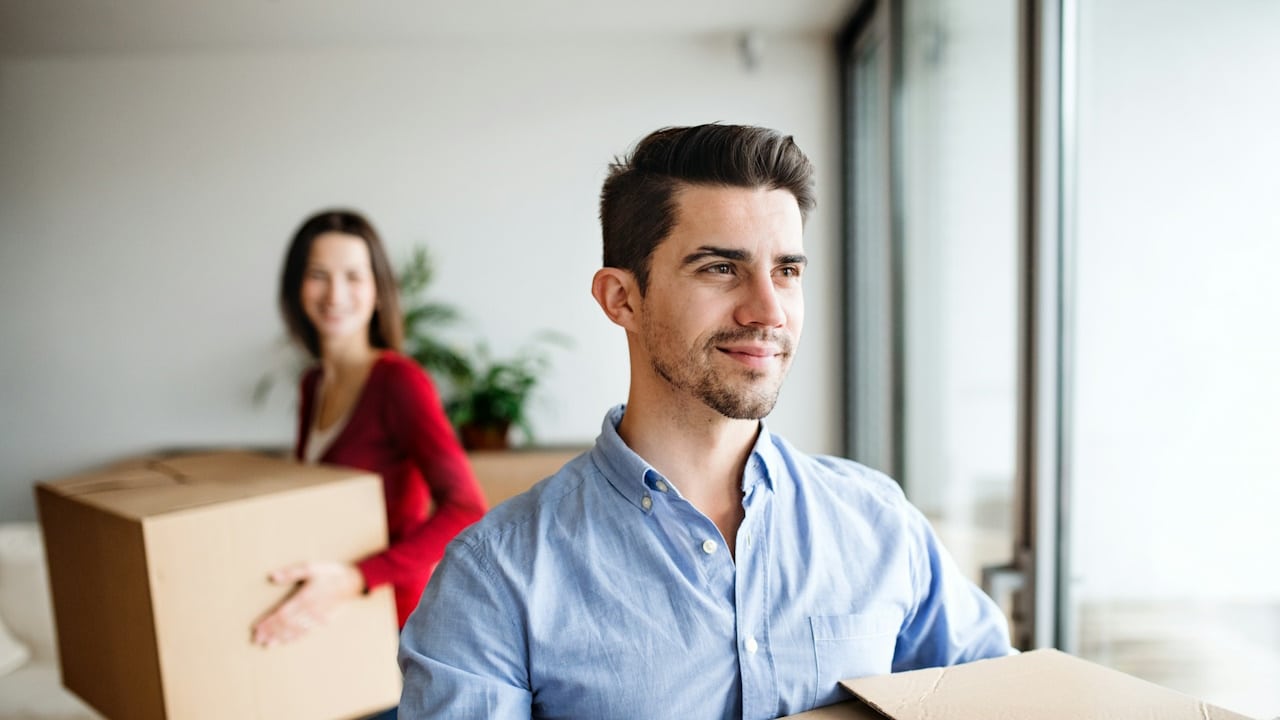 A man smiling and carrying a box as a woman in the background looks at him while carrying a box.