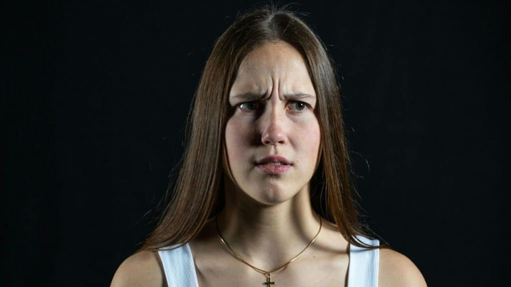 A young woman with long hair frowns, looking confused or upset, against a dark background.