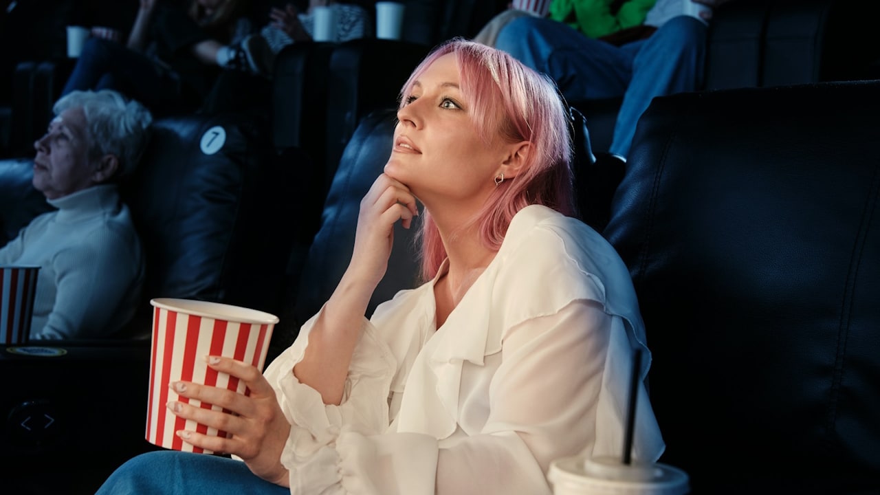 A young woman with pink hair, holding a tub of popcorn and watching a movie intently in a darkened theater.