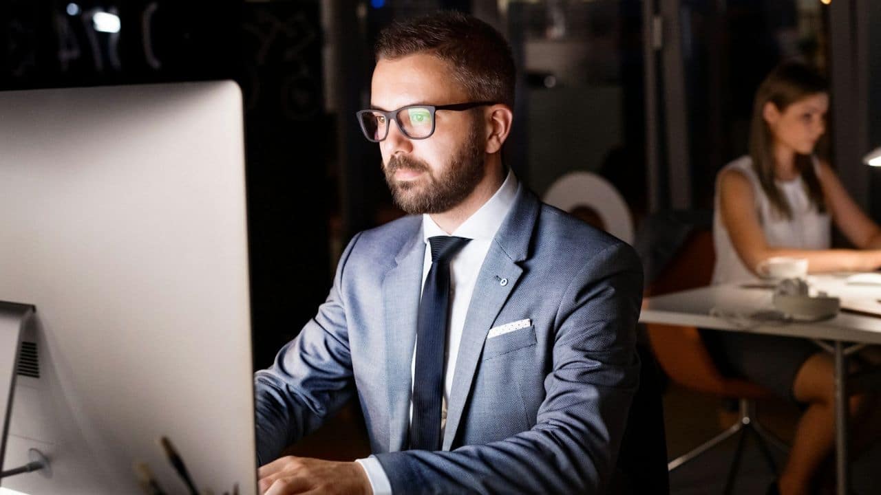 A businessman wearing glasses works late on a desktop computer.
