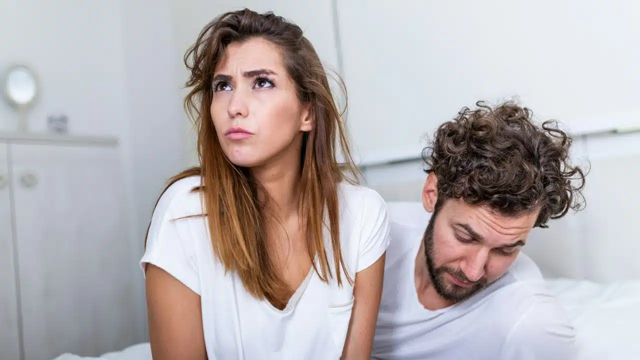 A distressed woman looks up while a man with curly hair bows his head behind her.