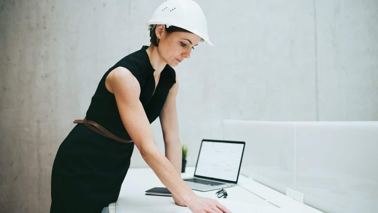 Woman in a white hard hat leaning over a desk with a laptop and papers.