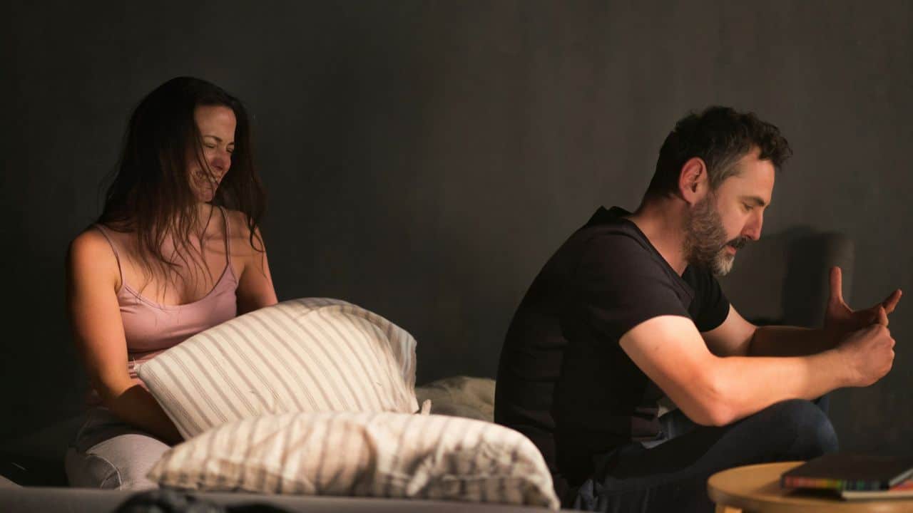 A man sits with his back to a crying woman holding a pillow on a bed in the dark.