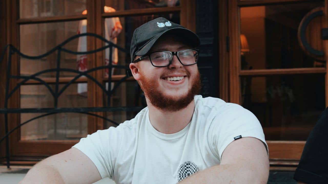 Smiling, bearded man in glasses and a black cap sitting outdoors, looking left.