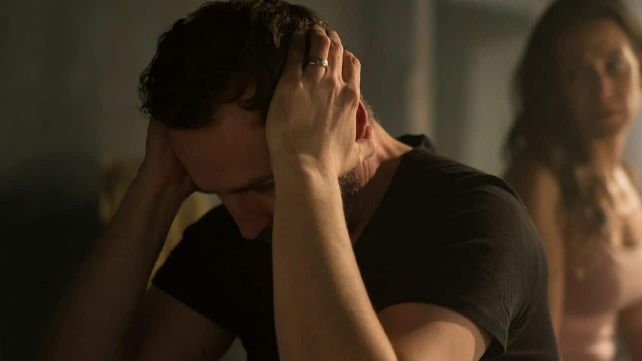 A man with short hair and a wedding ring holds his head in a dark room.
