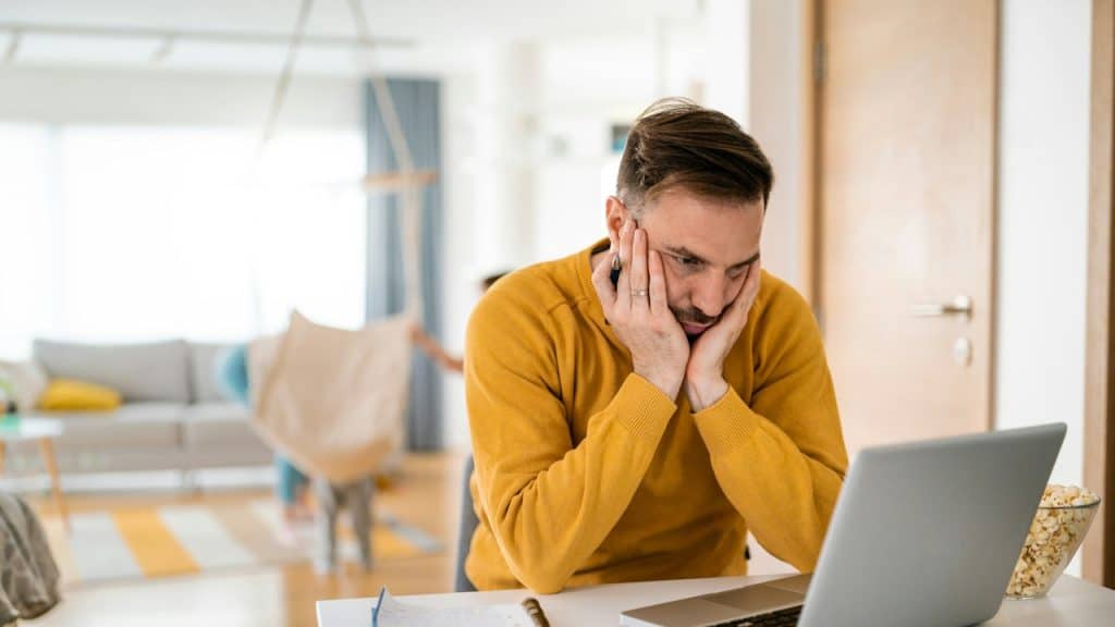 A tired man holding his face in his hands is working on the laptop.