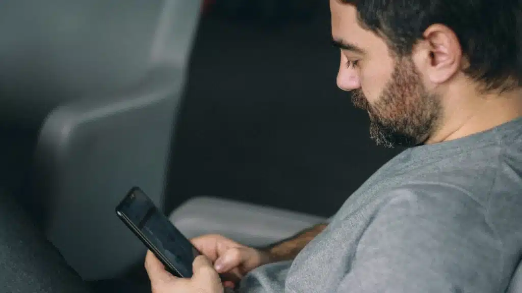 A bearded man sitting and looking at his phone’s screen.