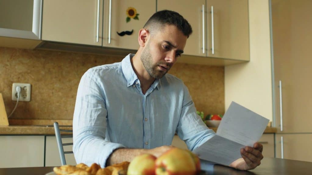 A man in a light blue shirt is sitting at a kitchen table reading a document.