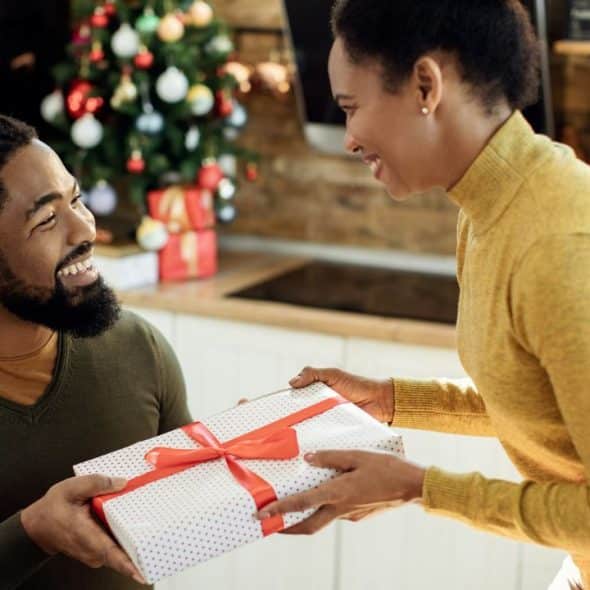 A woman in a yellow turtleneck is handing a Christmas present with a red ribbon to a smiling man.