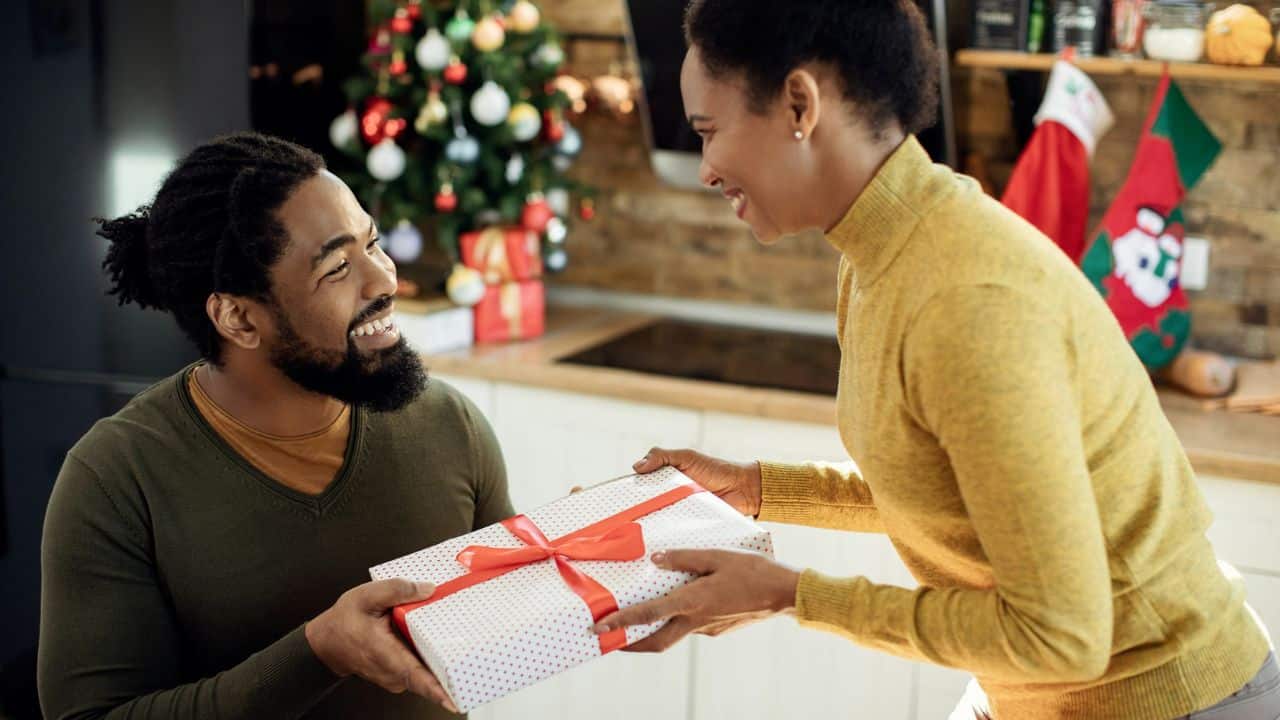 A woman in a yellow turtleneck is handing a Christmas present with a red ribbon to a smiling man.