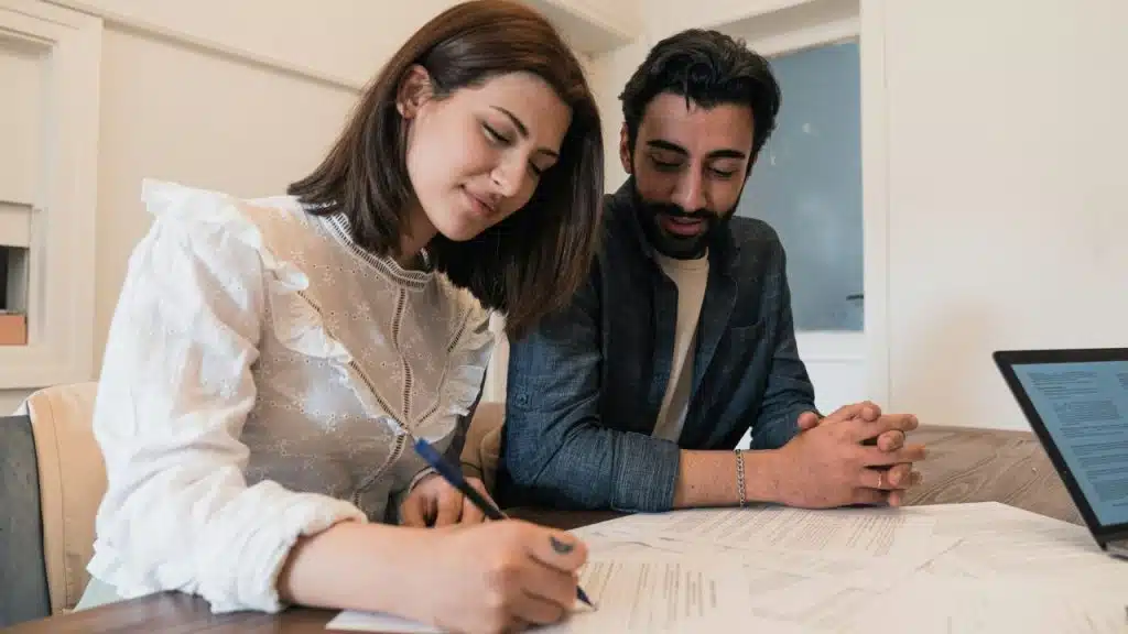 A woman signs documents while a man sits beside her with hands clasped on the table.