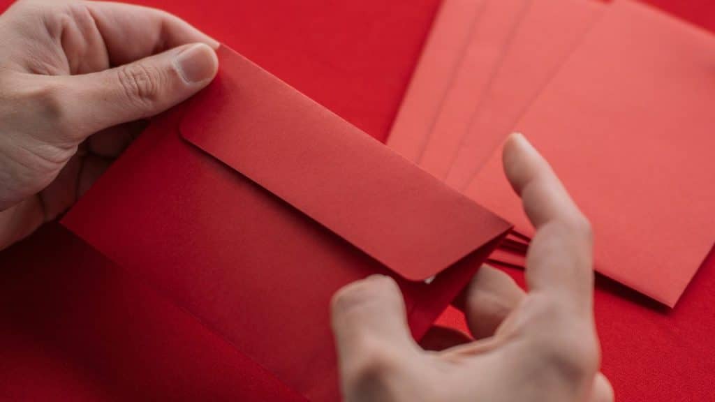 Hands hold and insert a gift card or paper into a red envelope on a red surface.
