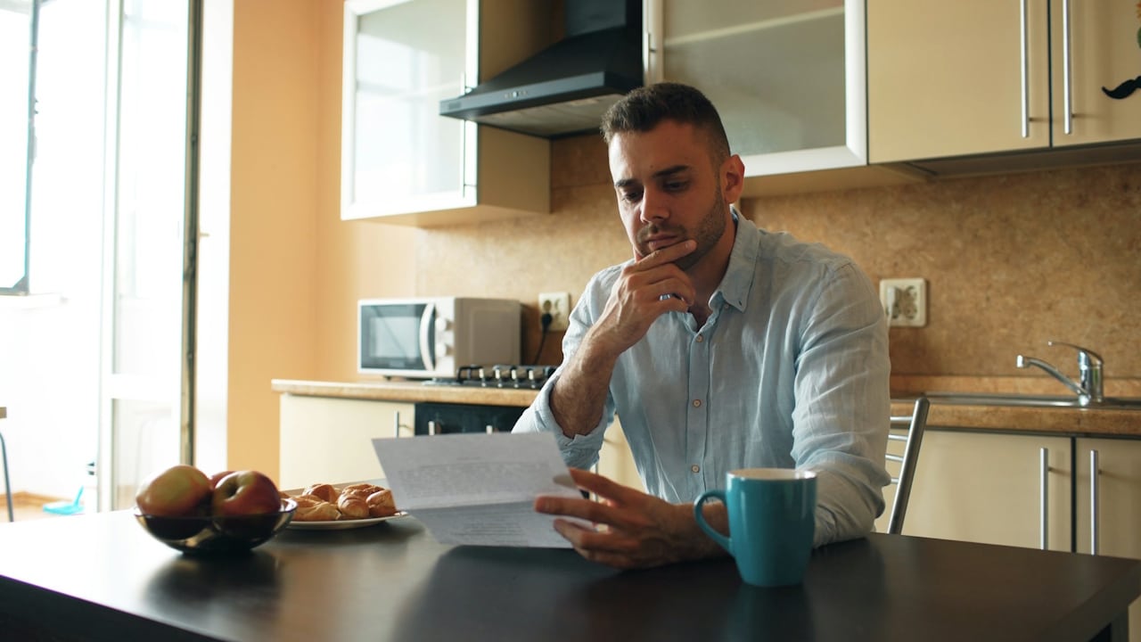 A man with a pensive expression reads a document while sitting at a kitchen table with a mug and a plate of food.