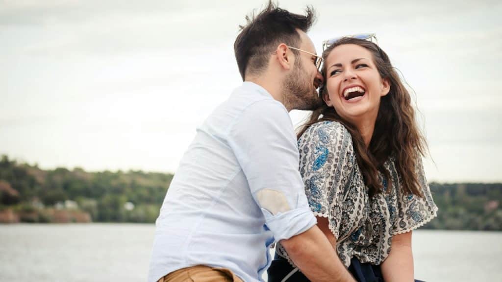 A lightly bearded man wearing sunglasses leans in closer to a laughing woman’s cheek to kiss her while they sit in front of a lake.