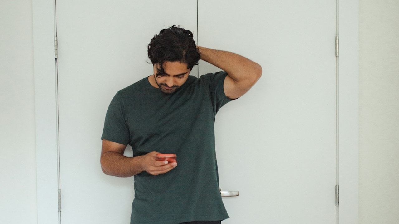 A man looking upset, holding his hand to the back of head and leaning against a door while looking at his phone’s screen.