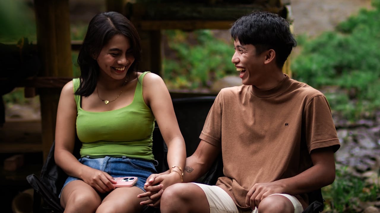 A smiling couple is sitting outdoors, holding hands and laughing together.
