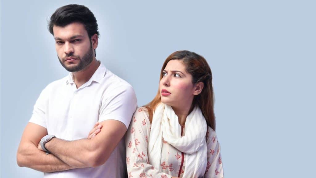 Man with crossed arms looking serious, while a woman looks at him cautiously.