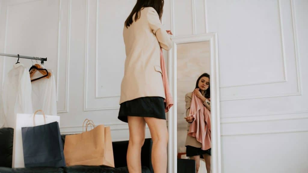 A woman in a blazer stands before a full-length mirror, looking at her reflection.