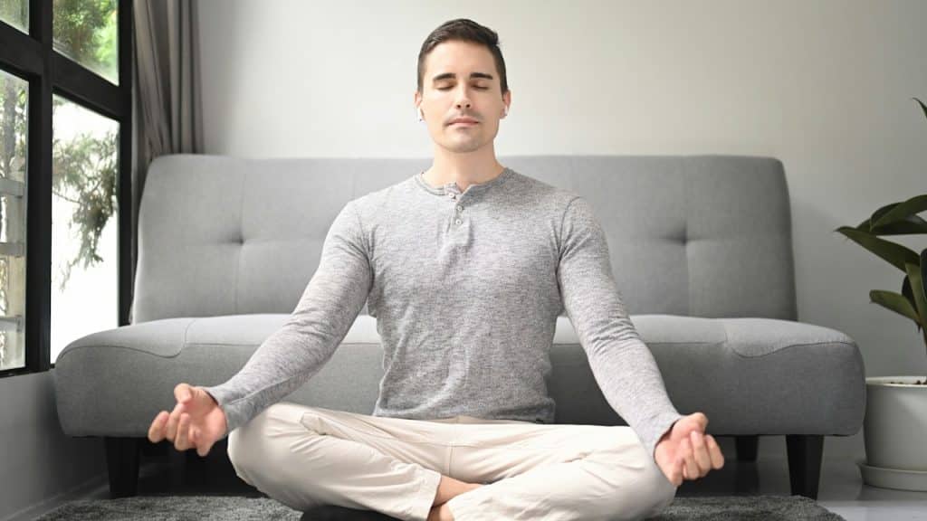 A man with his eyes closed is sitting in a lotus position on a rug in a living room, meditating.