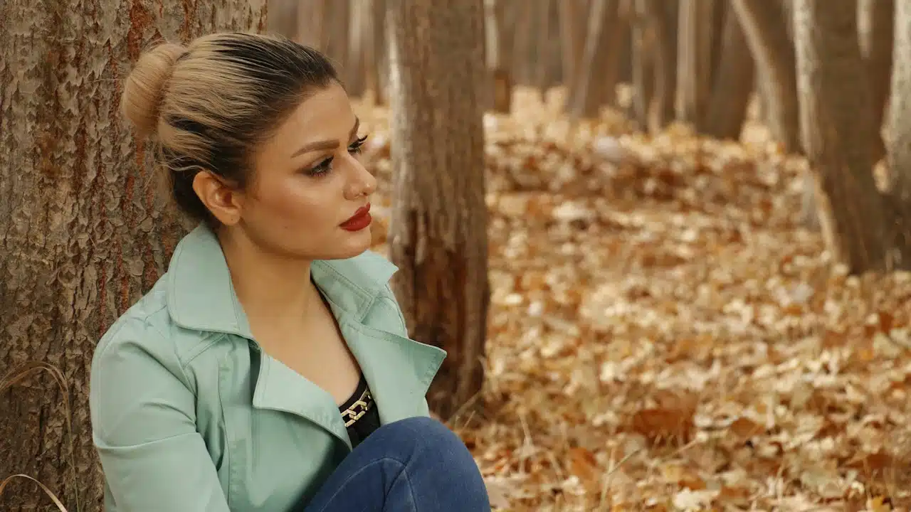 A woman is sitting on the ground next to a tree in fall.