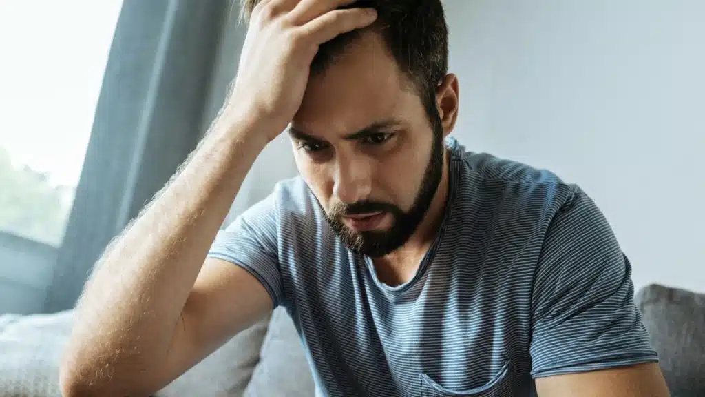 Close-up of a man with a beard and striped shirt, clutching his forehead in distress.