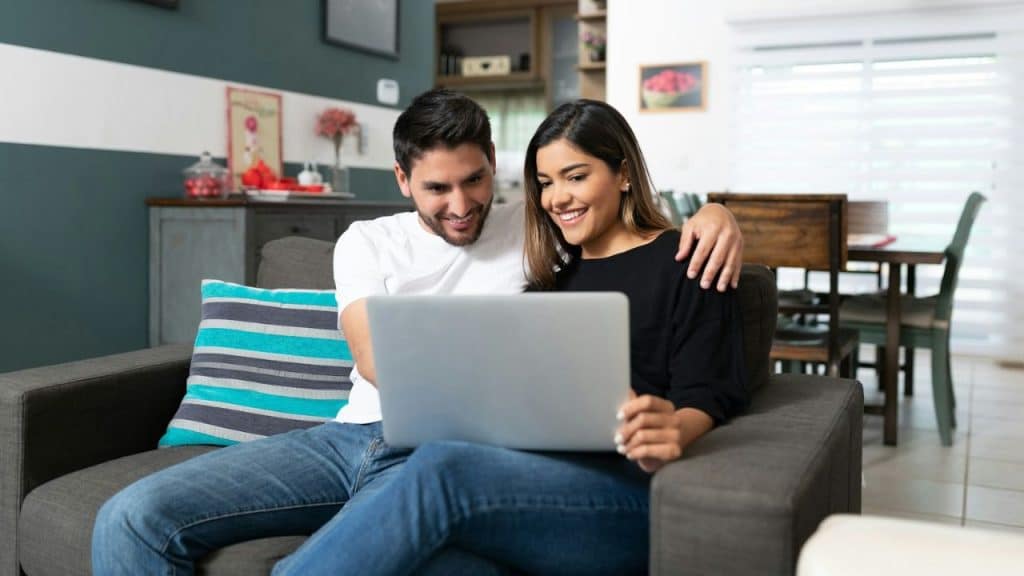A smiling couple sits on a gray sofa looking at a laptop together.
